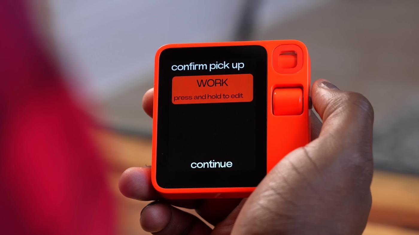A hand holding the Rabbit r1, an orange pocket AI device, showing its screen reading confirm pick up, WORK, press and hold to edit, continue — a real photograph.