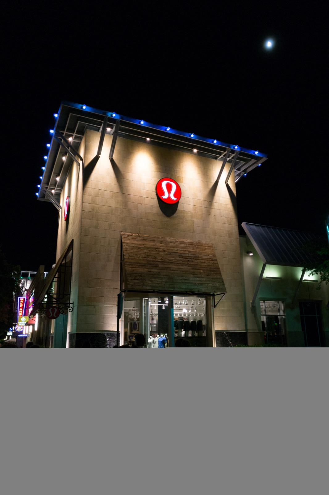 A Lululemon store exterior at night, illuminated storefront with the brand's logo, a real photograph.