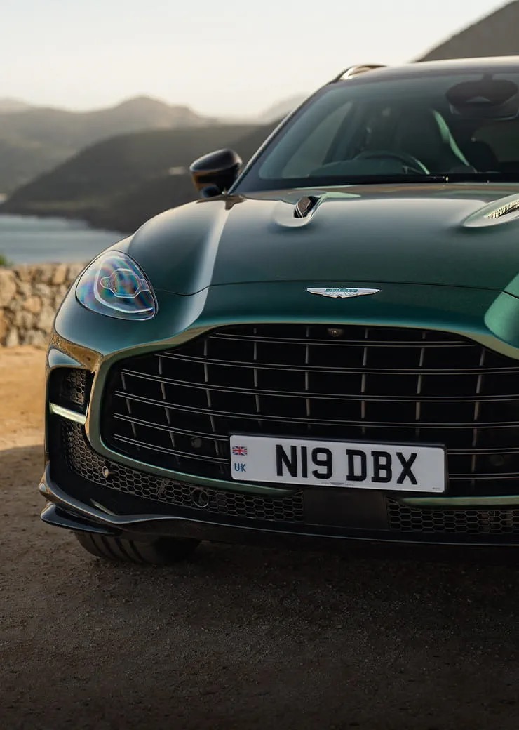 Close-up front view of the Aston Martin DBX707's grille and headlight, parked overlooking a coastline.