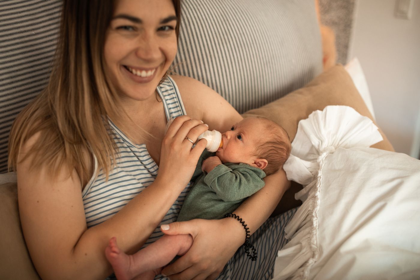 A smiling parent bottle-feeding a newborn while seated on a couch