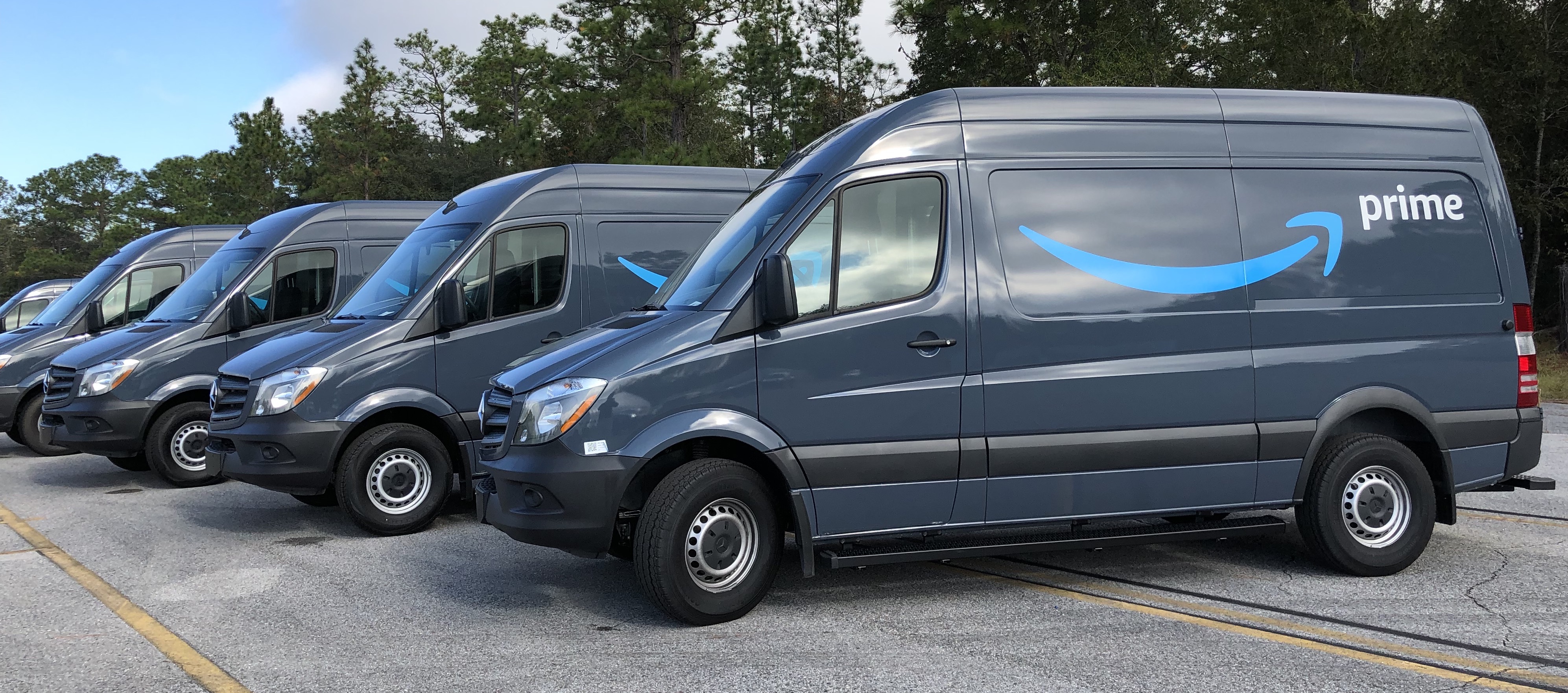 A row of Amazon Prime-branded delivery vans parked in a lot, real photograph.