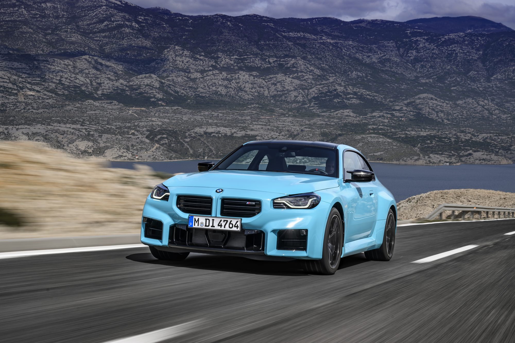 BMW M2 with M xDrive in light blue, photographed in motion on a coastal mountain road.