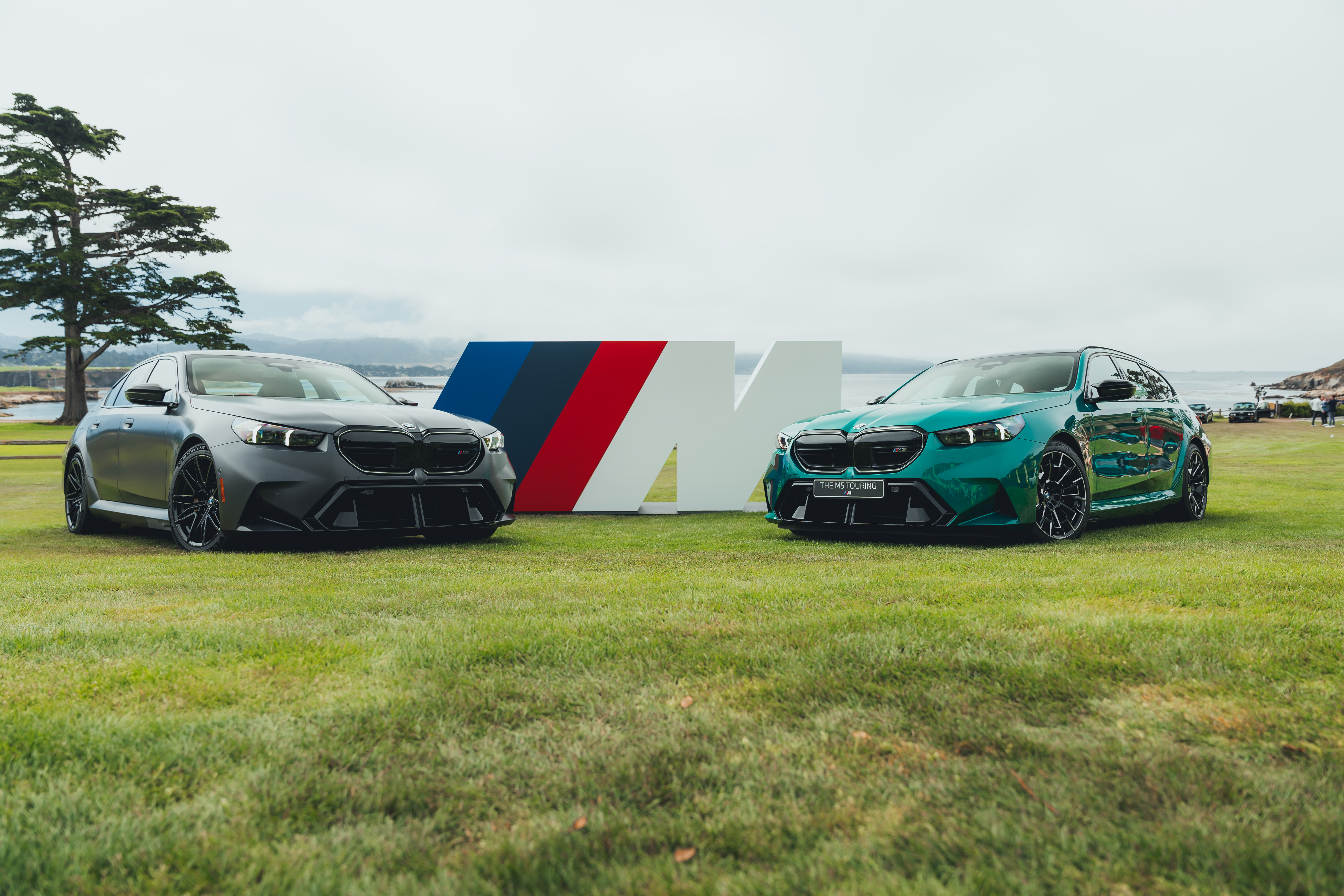 A grey BMW M5 sedan and a green BMW M5 Touring parked together on grass, with an M-logo sign between them, at the model's world premiere event.