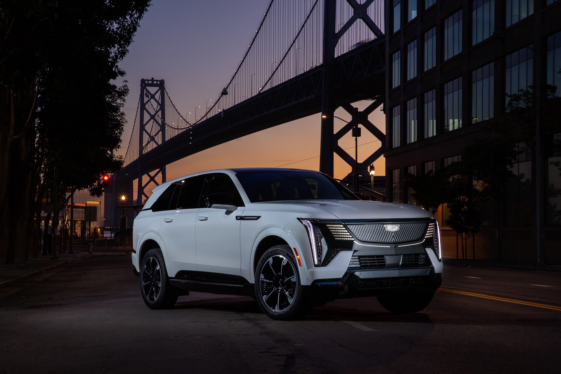 A white Cadillac Escalade IQ parked on a city street at dusk with a suspension bridge behind it.