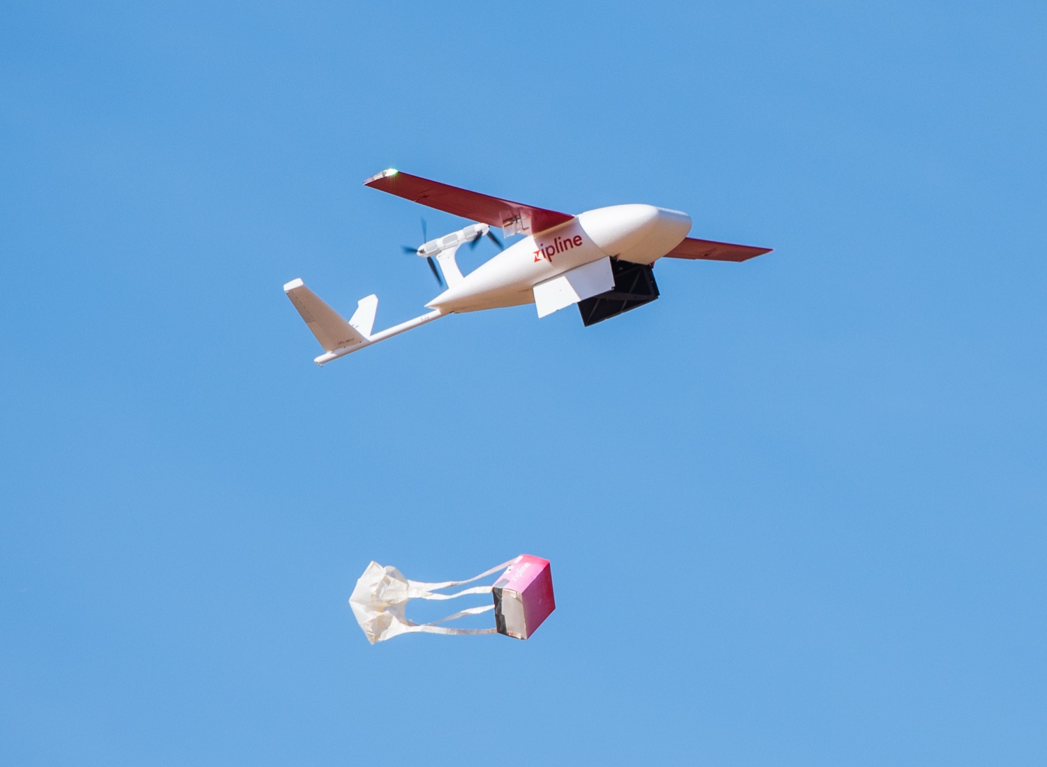 A Zipline delivery drone releasing a package by parachute in flight.