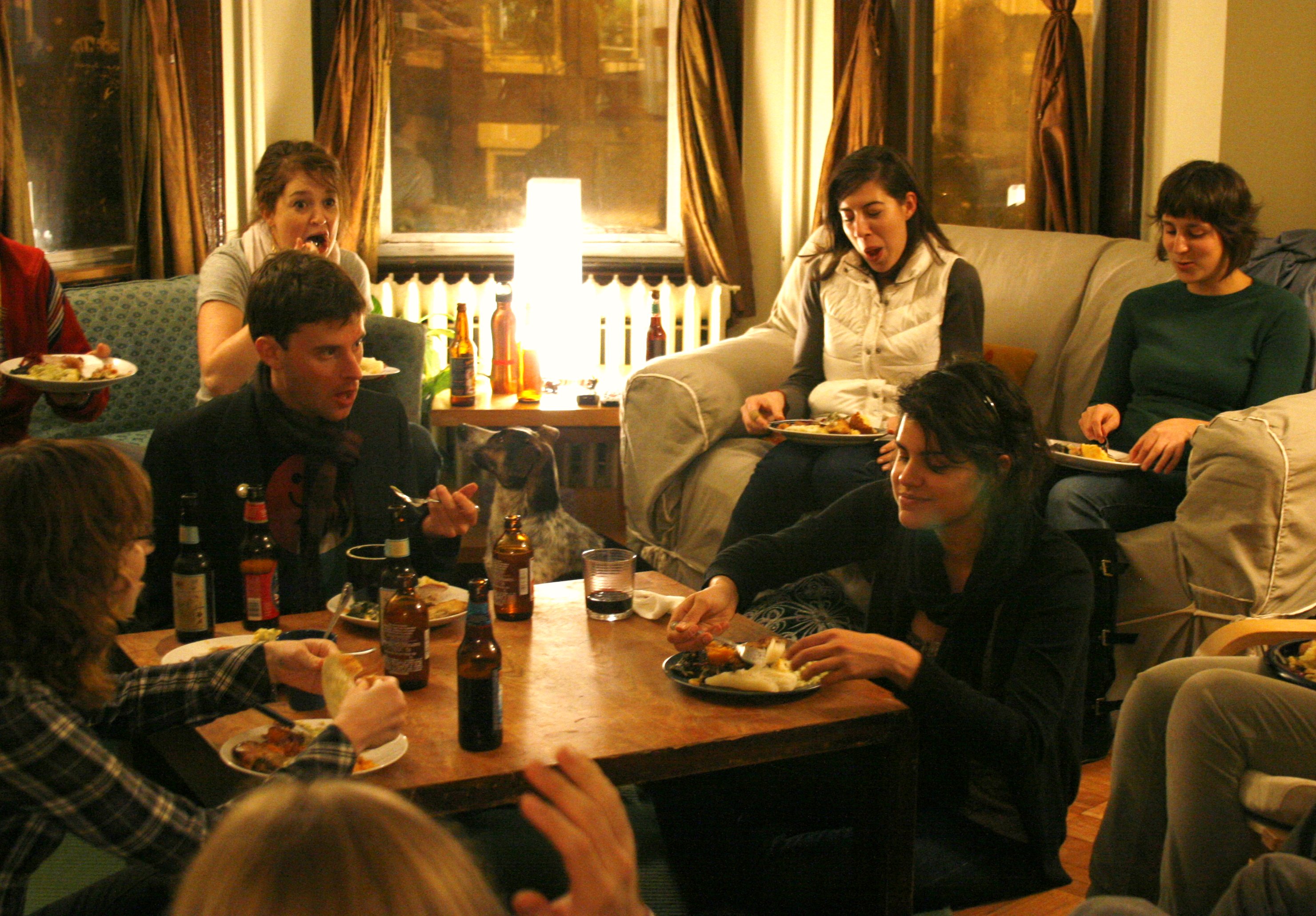 A group of friends eating a casual potluck meal together in a living room, real photograph.