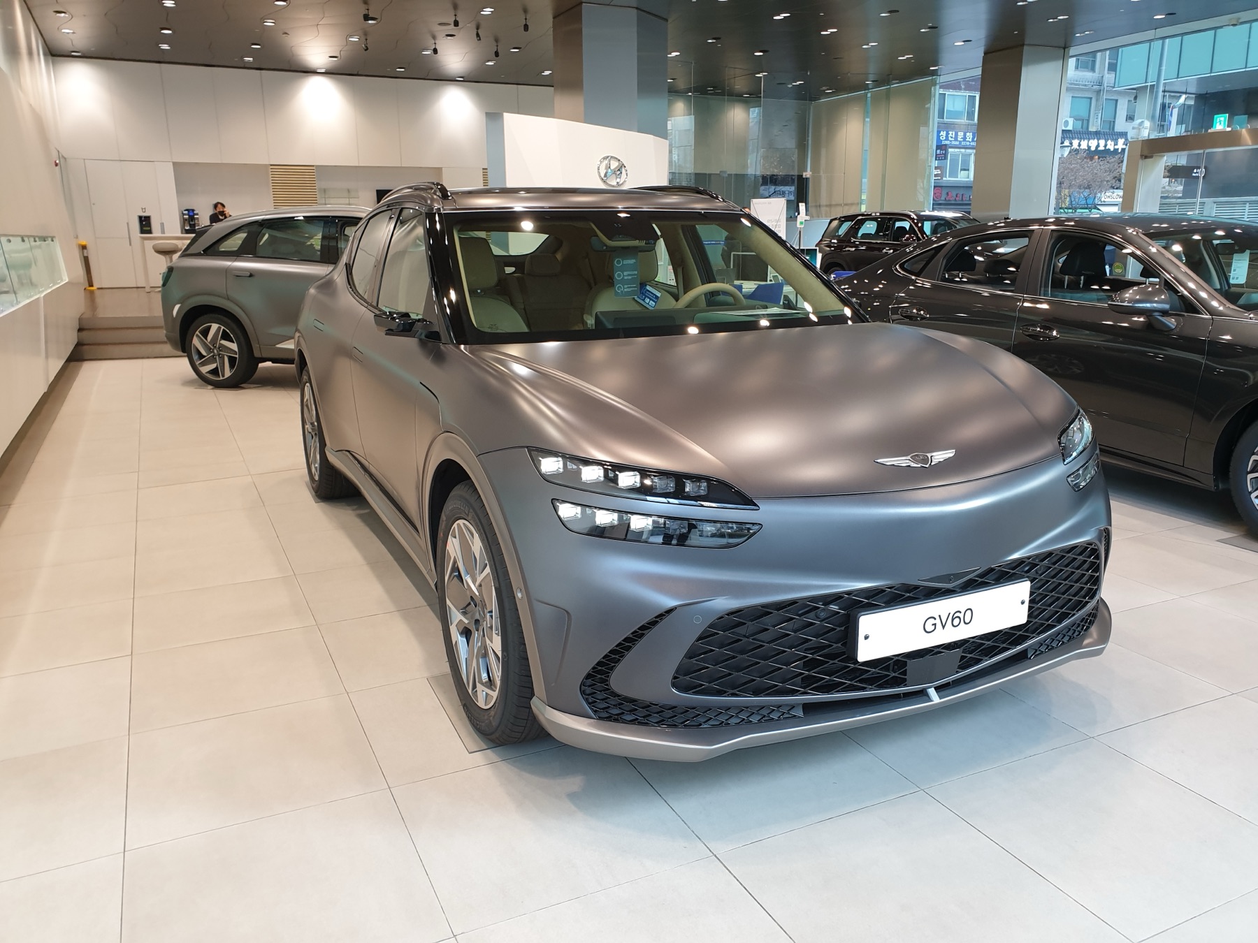 Genesis GV60 in a matte grey finish, front three-quarter view, dealership showroom.