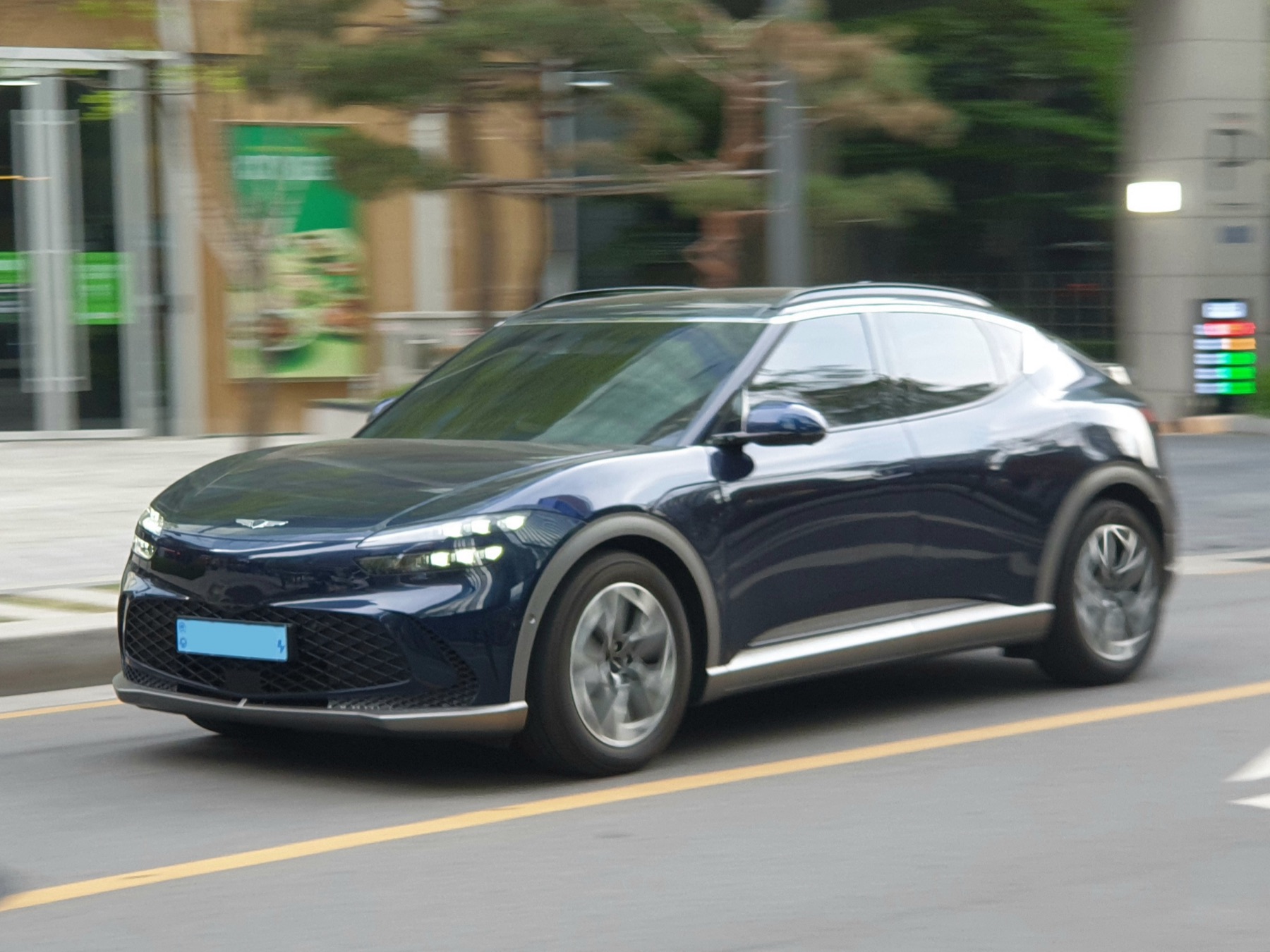 Genesis GV60 front three-quarter view in dark blue, driving on a city street, real-world photography.