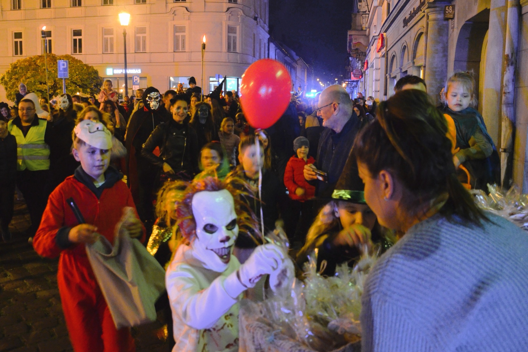 A nighttime Halloween parade crowd in costume, including a clown-style mask and a red balloon, a real photograph.