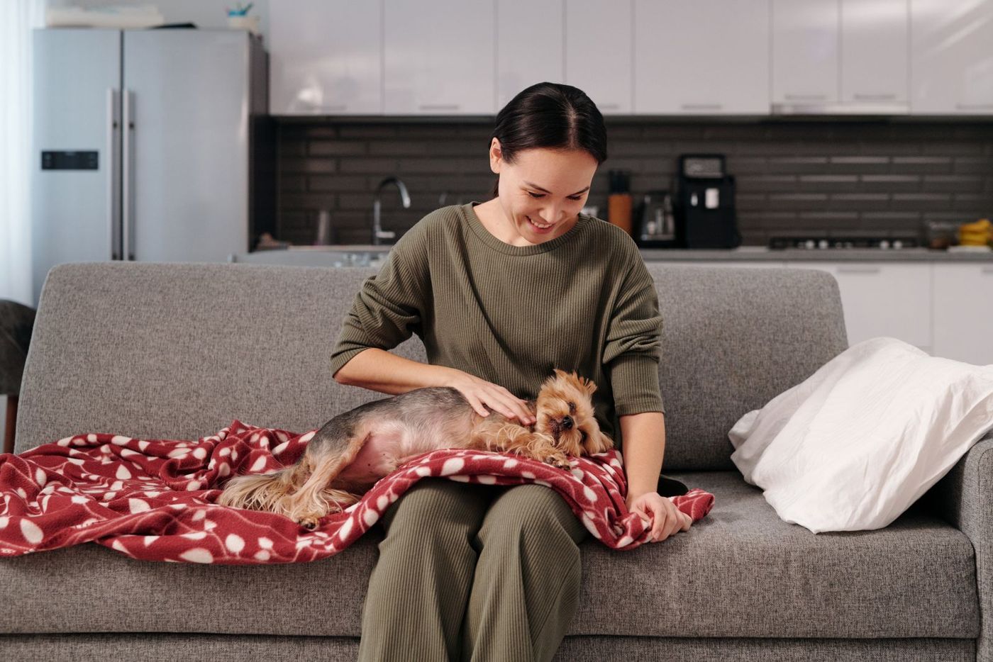 A woman smiling and petting a small dog resting on a red blanket on a sofa