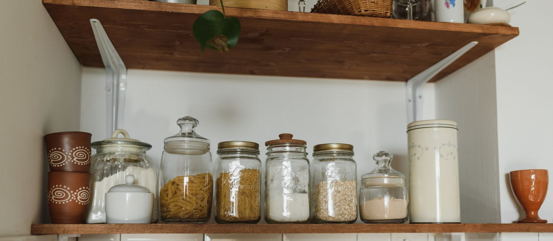 A wooden kitchen shelf holding glass jars of pasta, flour and oats.