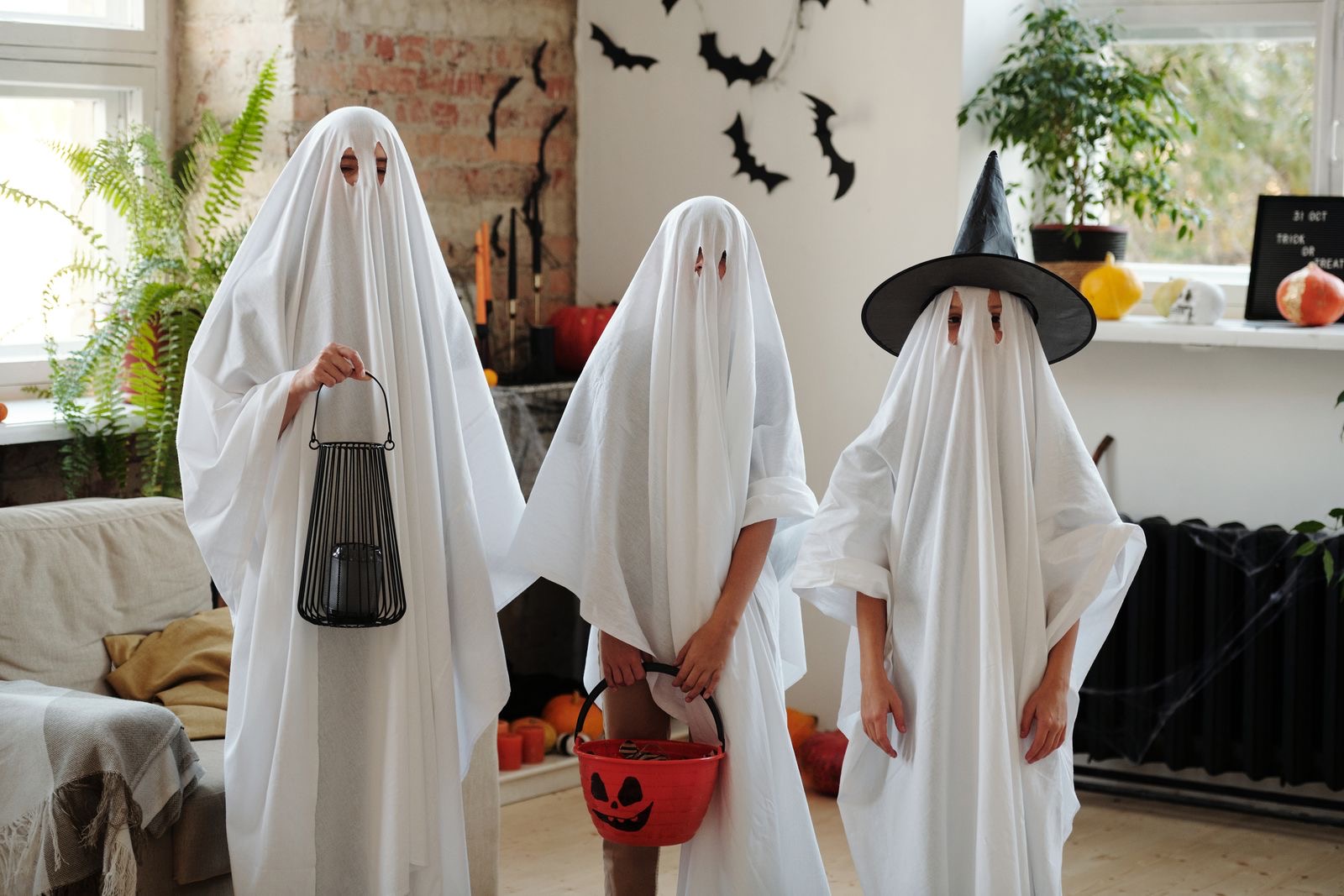 Three people in homemade white-sheet ghost costumes in a living room with Halloween decorations, one wearing a witch hat over the sheet.