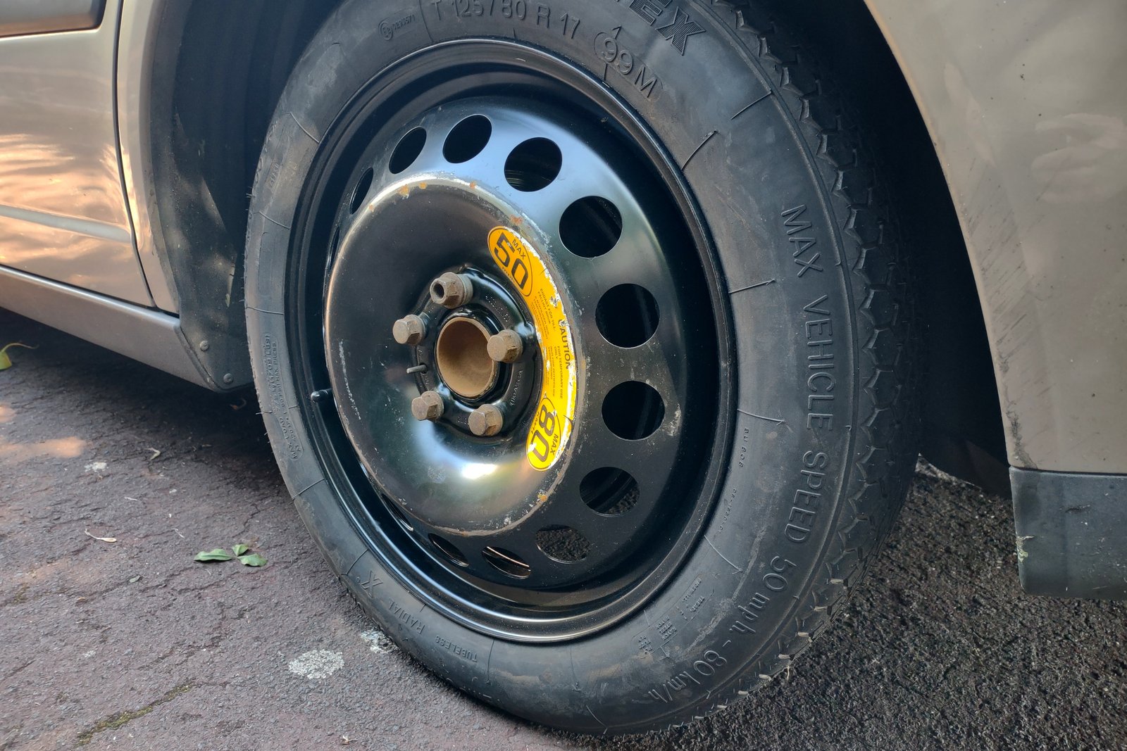 A compact spare tire correctly mounted and secured on the wheel hub, with its temporary-use speed rating label visible