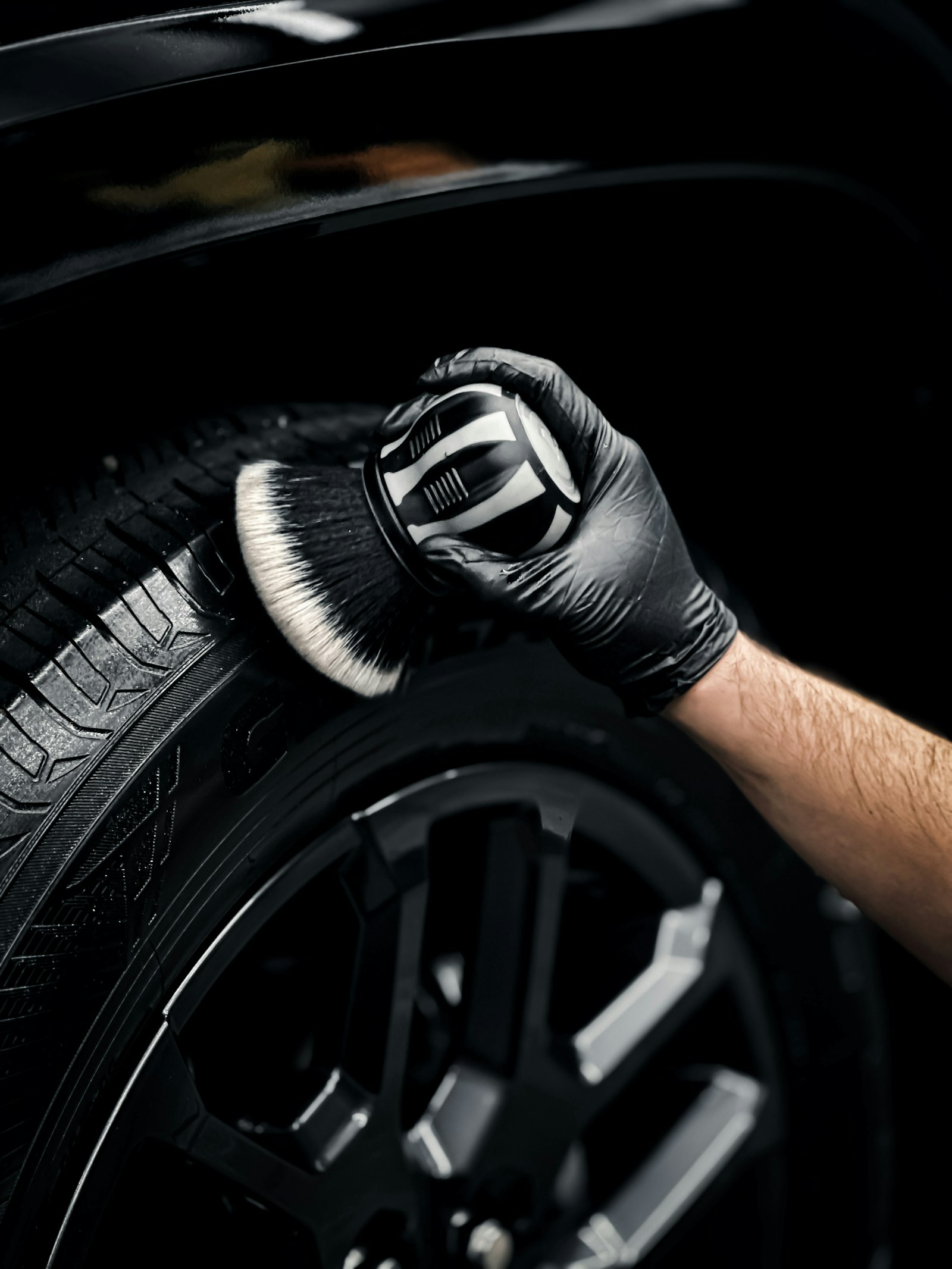 Gloved hands cleaning a car tire and wheel area with a detailing brush.