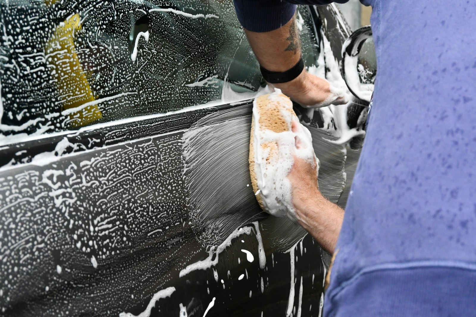 A person washing a car exterior with a sponge, working section by section.