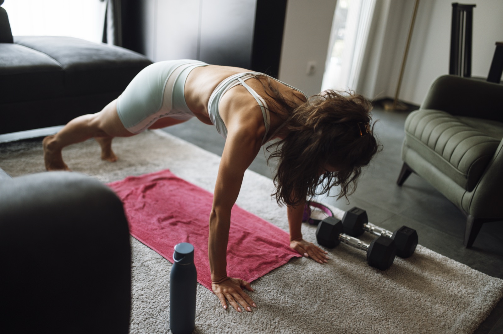 A woman doing a push-up on a towel next to a set of dumbbells in a small living room workout area.
