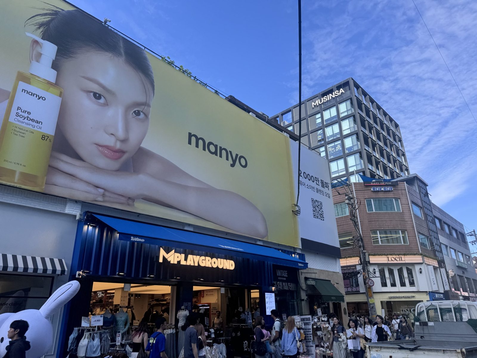 A Seongsu street corner under a large Manyo billboard, with shoppers outside the MPlayground store.