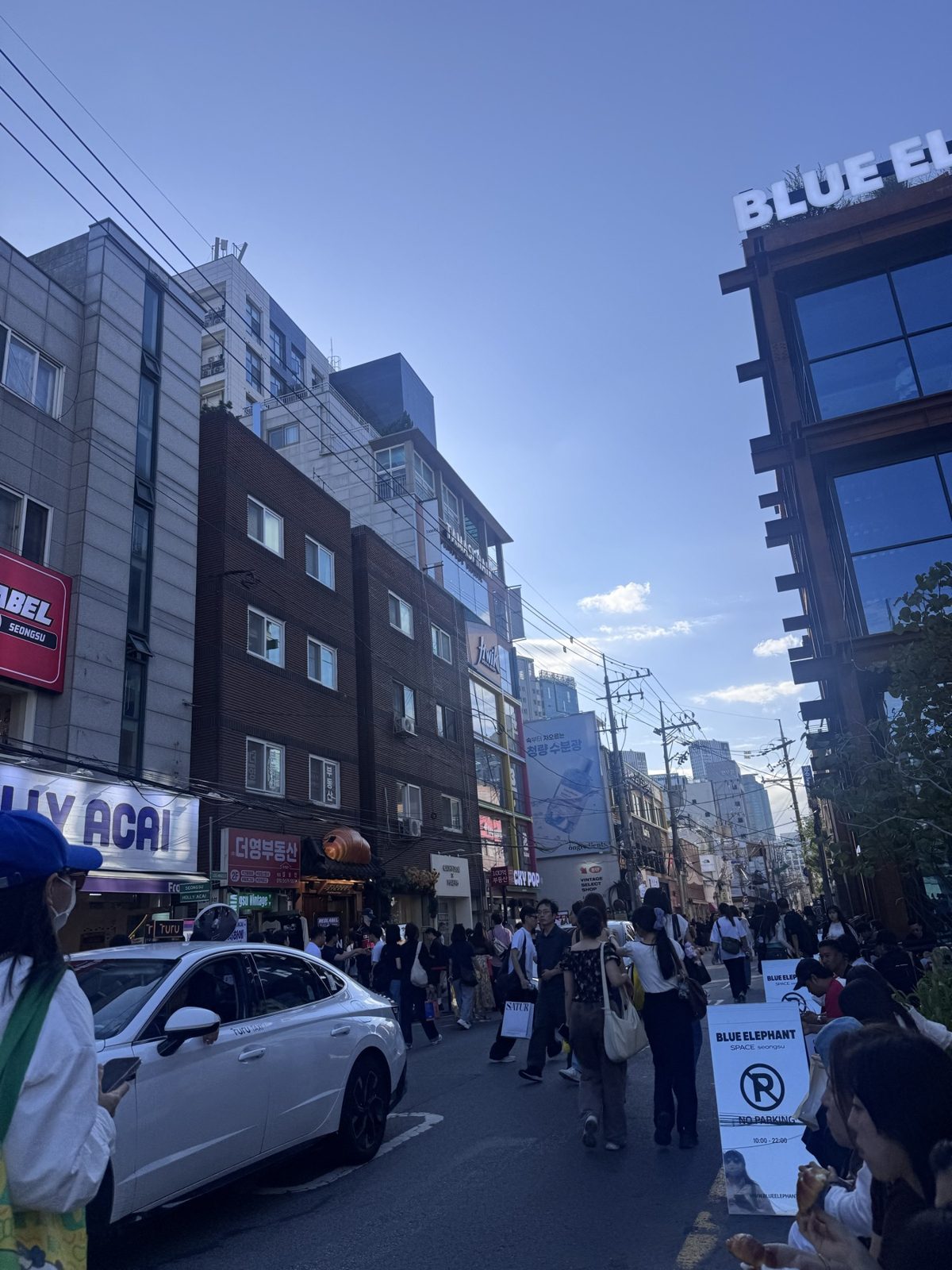 A busy Seongsu street on a clear afternoon, crowds along the road between shops and mid-rise buildings.