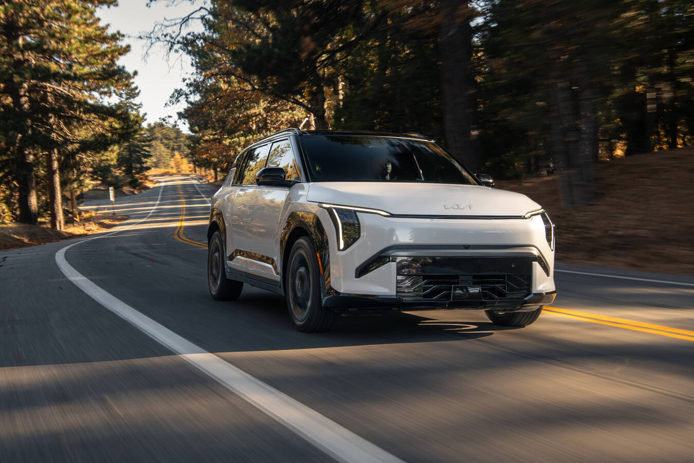 2027 Kia EV3 driving on a forested mountain road, front three-quarter view, official Kia press photography.
