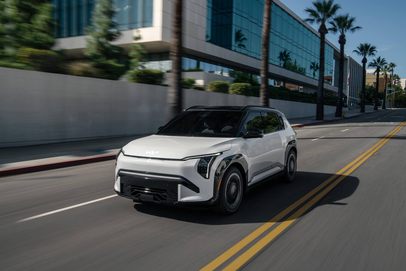 2027 Kia EV3 driving on a city street with palm trees, front three-quarter view, official Kia press photography.
