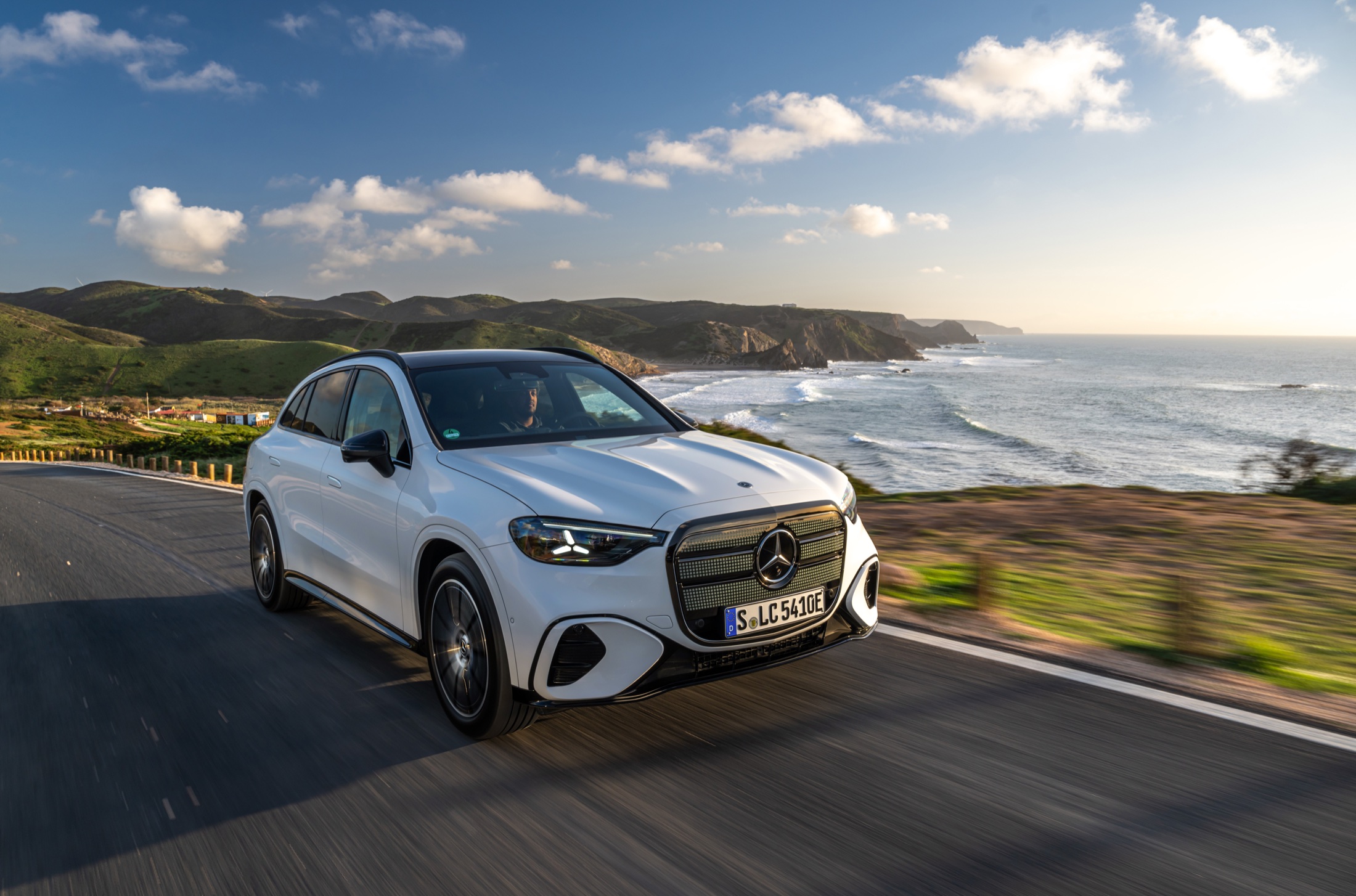 A white electric Mercedes GLC driving on a coastal road at golden hour, three-quarter front view with the illuminated grille lit, ocean and headland behind.