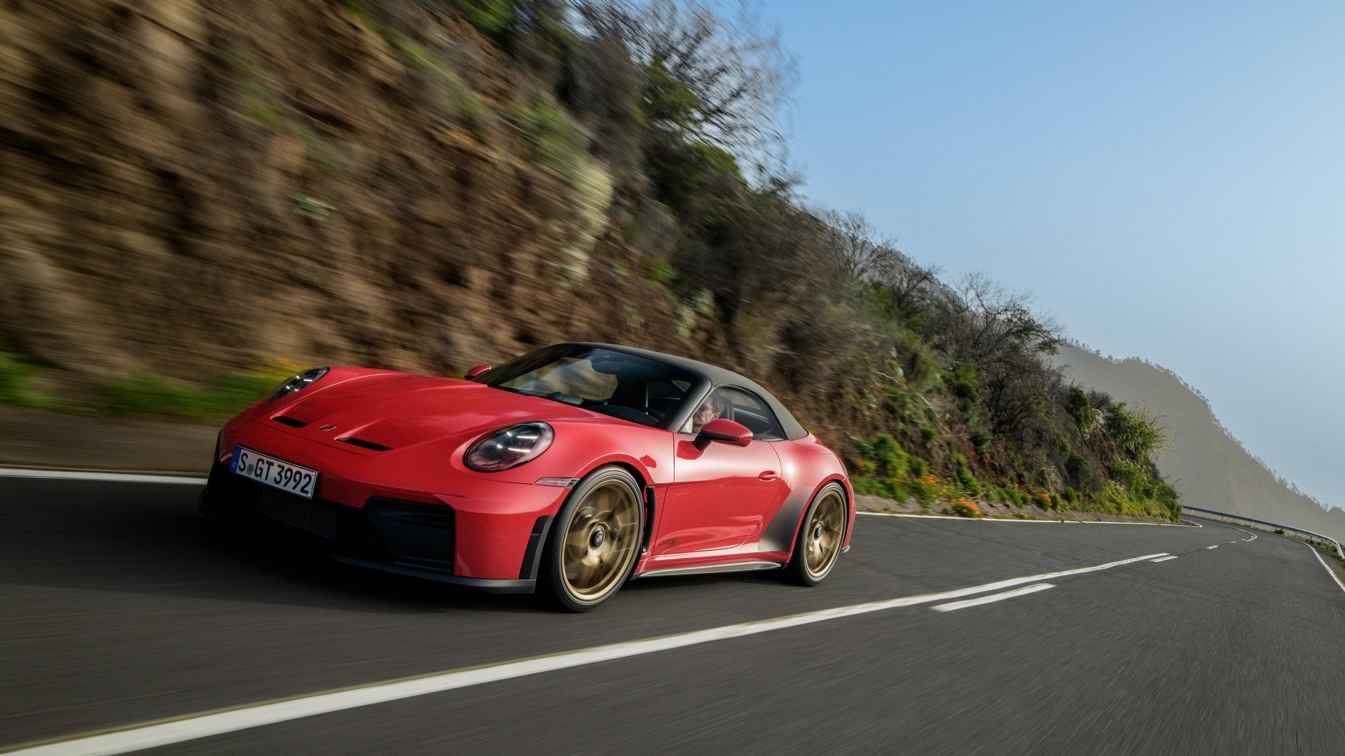 A red Porsche 911 GT3 S/C with the roof down, driving on a mountain road, front three-quarter view.