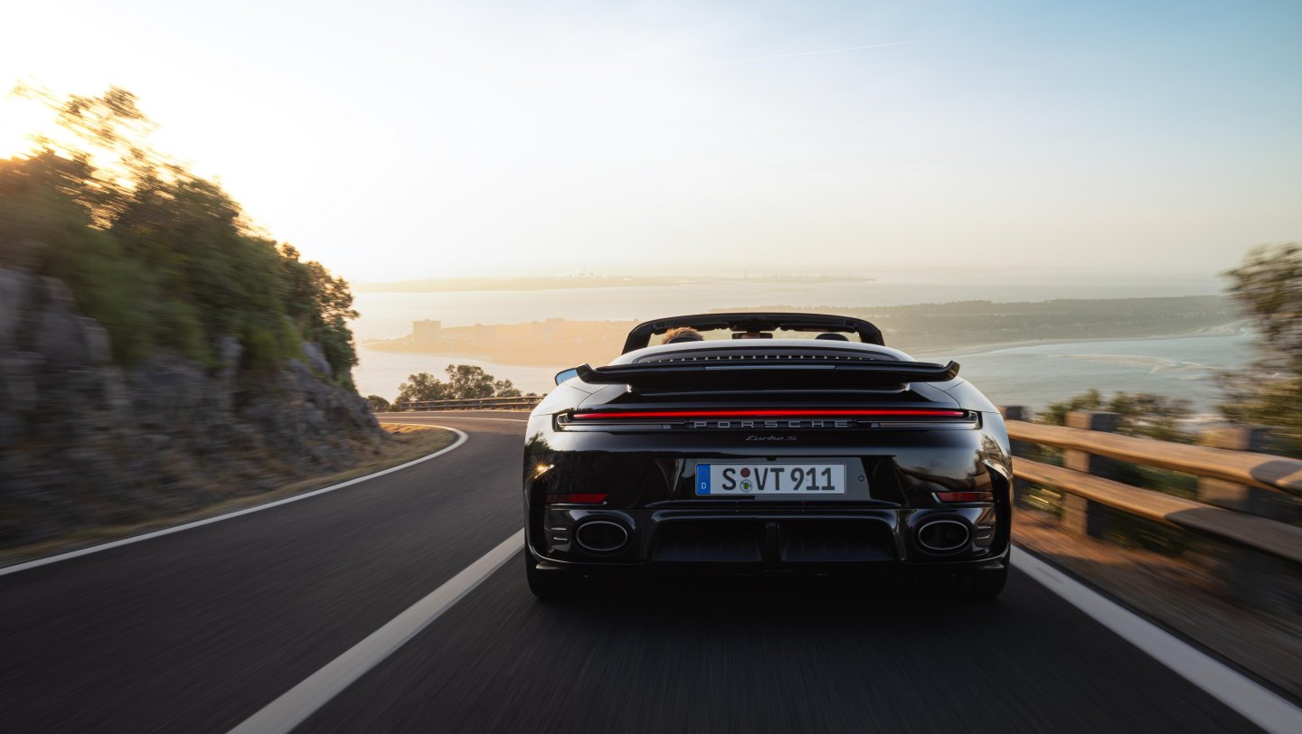 Rear view of the 911 Turbo S Cabriolet driving along a coastal road at sunset.