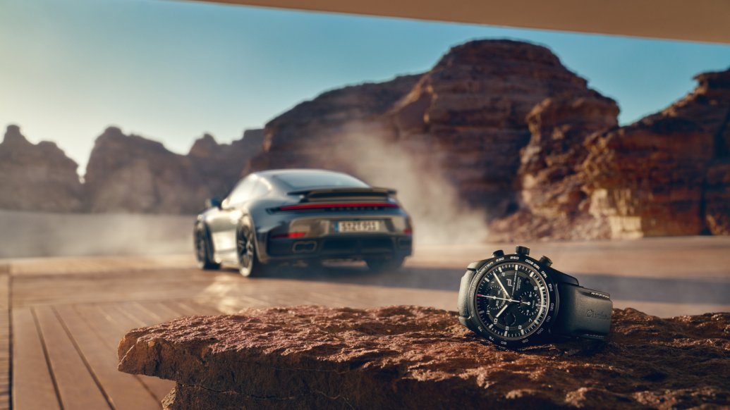 911 Turbo S Coupe drifting on a desert road, kicking up dust, seen from behind a foreground rock.
