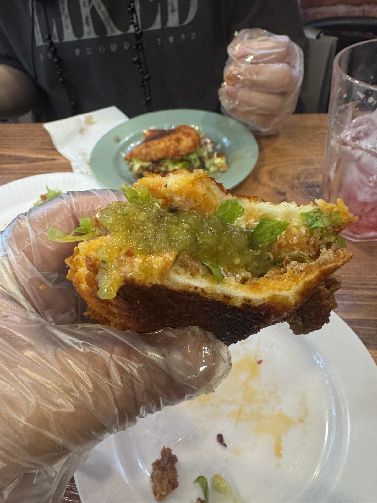 A pambazo held in a gloved hand, cut open to show the saucy bread, filling and green salsa.