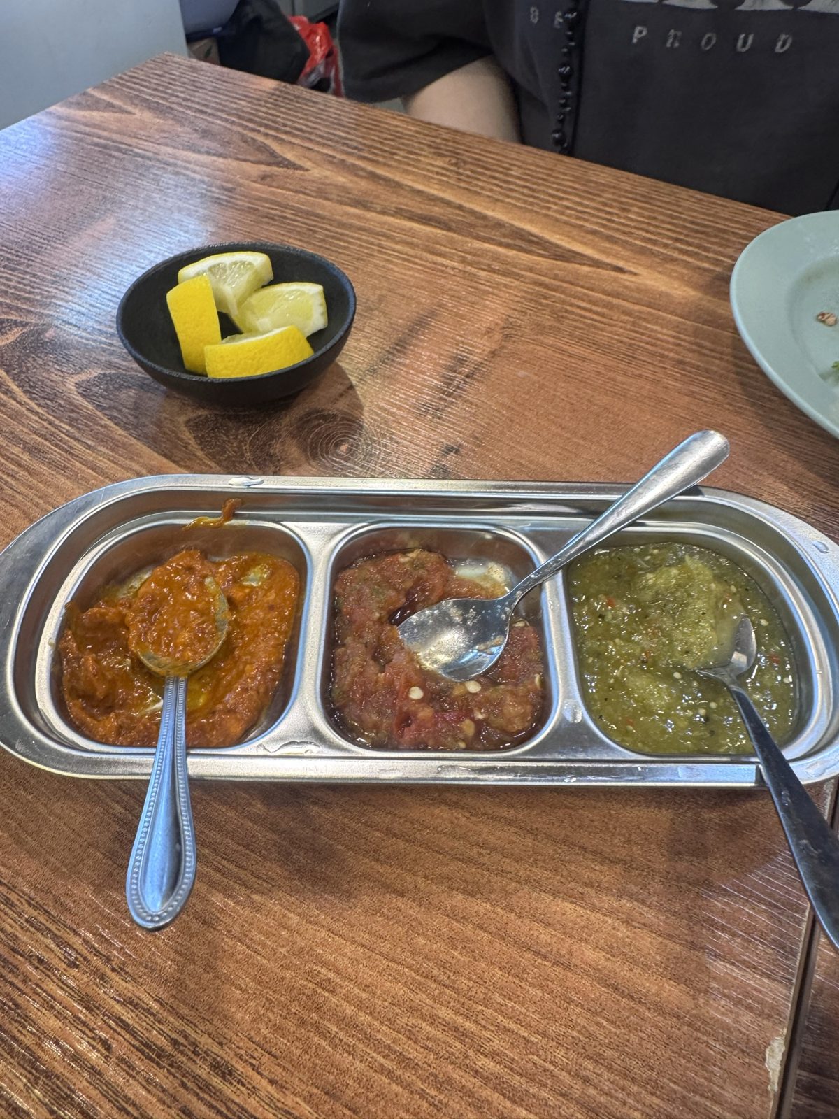 A three-compartment metal tray of salsas — red, dark red and green — with spoons, and lemon wedges behind.
