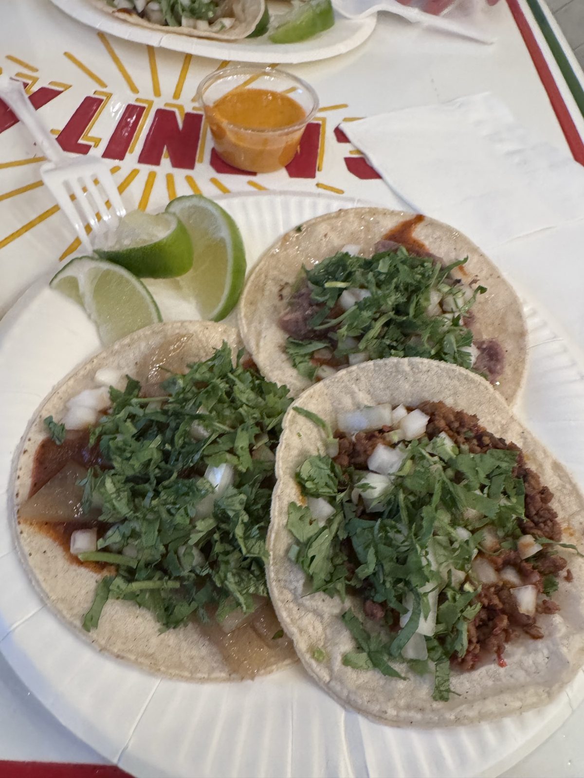 Three tacos at Villa Guerrero topped with onion and cilantro, lime wedges on the side, on a disposable plate.
