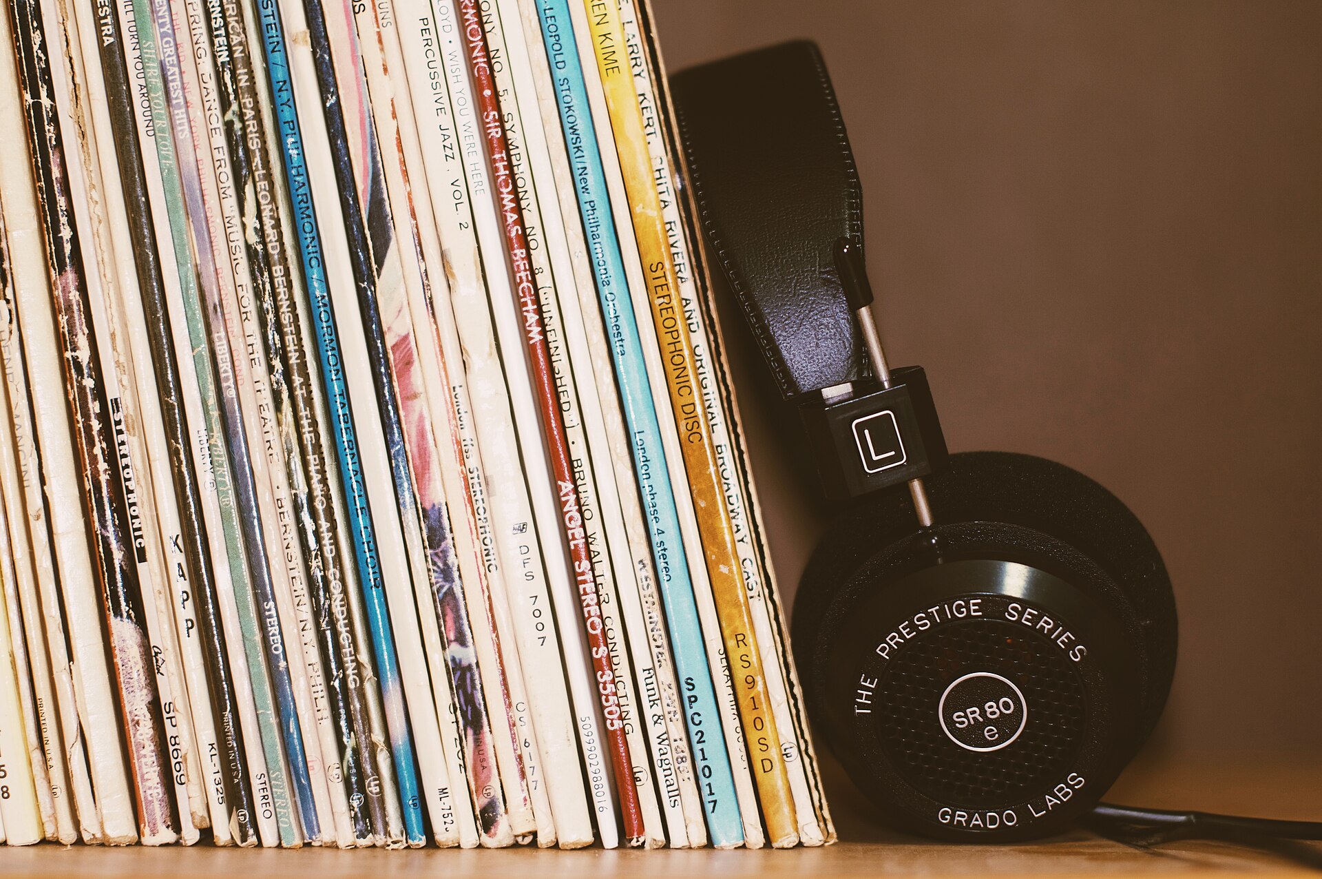 A stack of vinyl record sleeves leaning against a pair of over-ear headphones on a wooden shelf, real photograph.