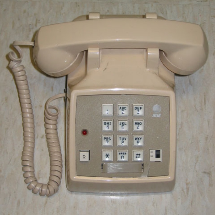 A cream-colored AT&T push-button telephone with a coiled cord, mounted on a wall — the exact kind of phone you'd have used to dial 777-FILM.