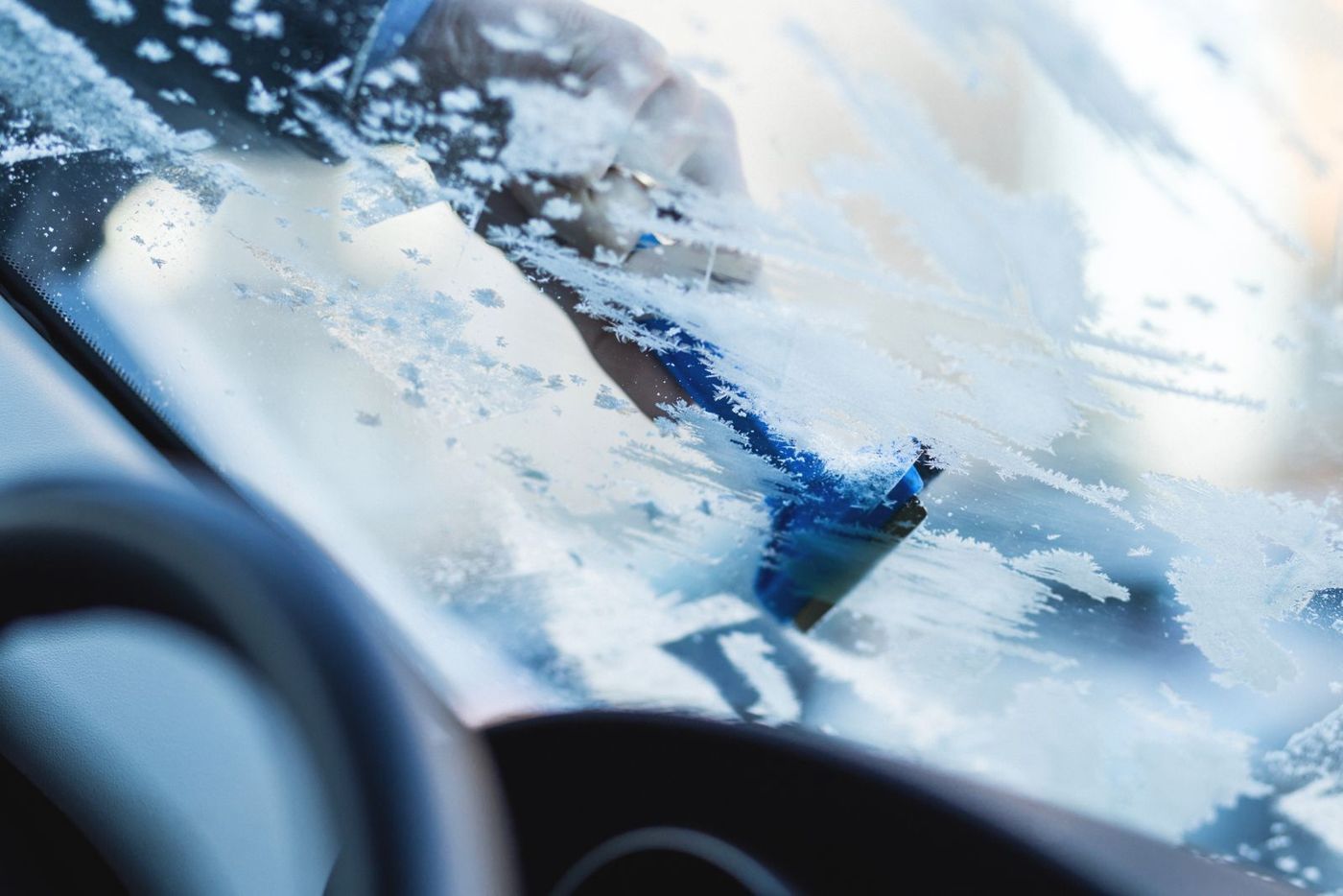 A hand scraping frost off the inside of a car windshield with a blue ice scraper
